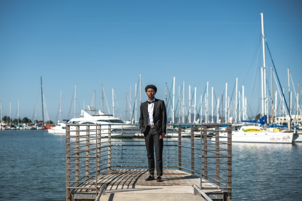 A young man stands at the end of a pier wearing a tuxedo with a backdrop of boats behind him during a senior photoshoot in Richmond, CA
