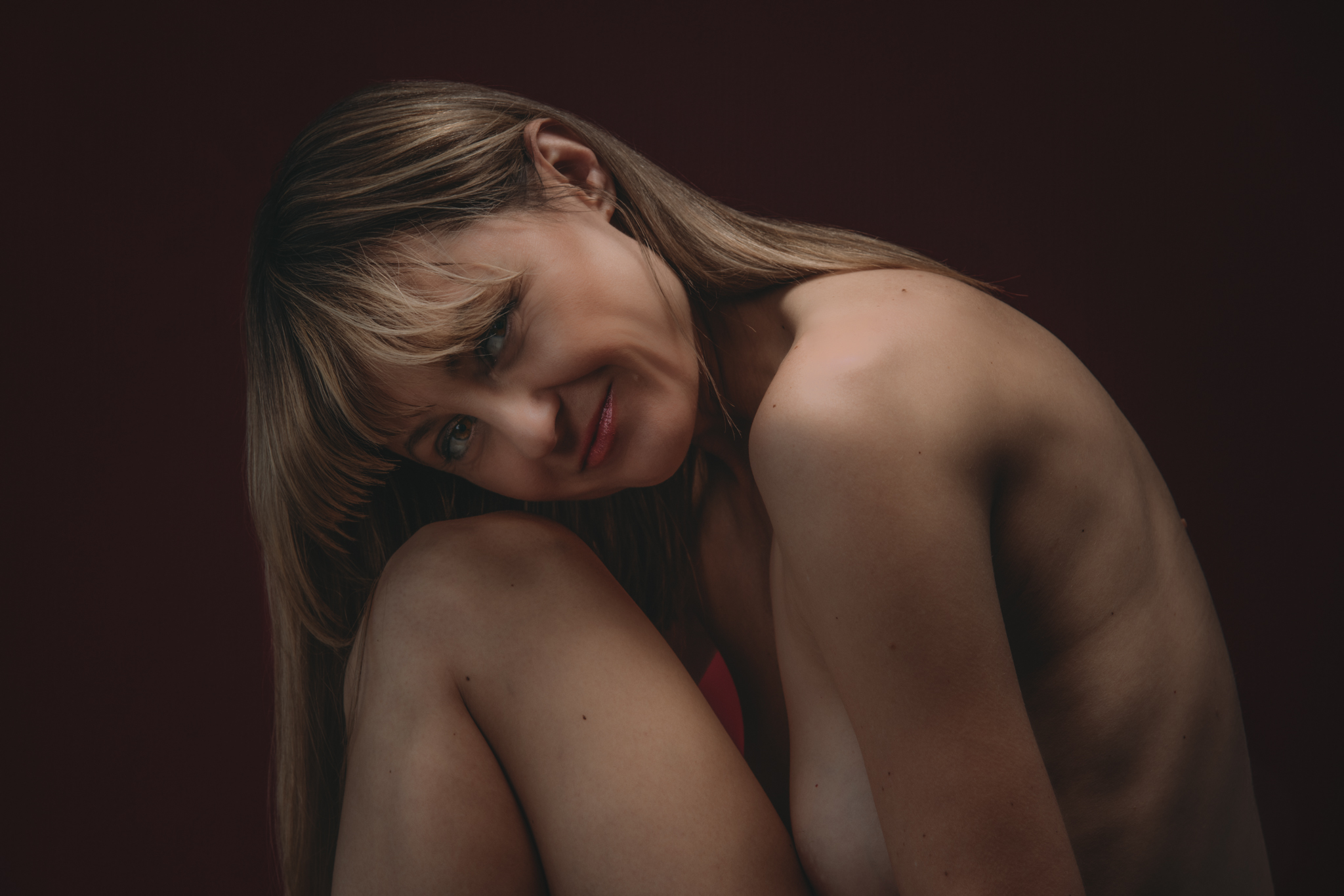 Woman lays her head on her knee against a dark red backdrop during a boudoir photo shoot in San Francisco, CA