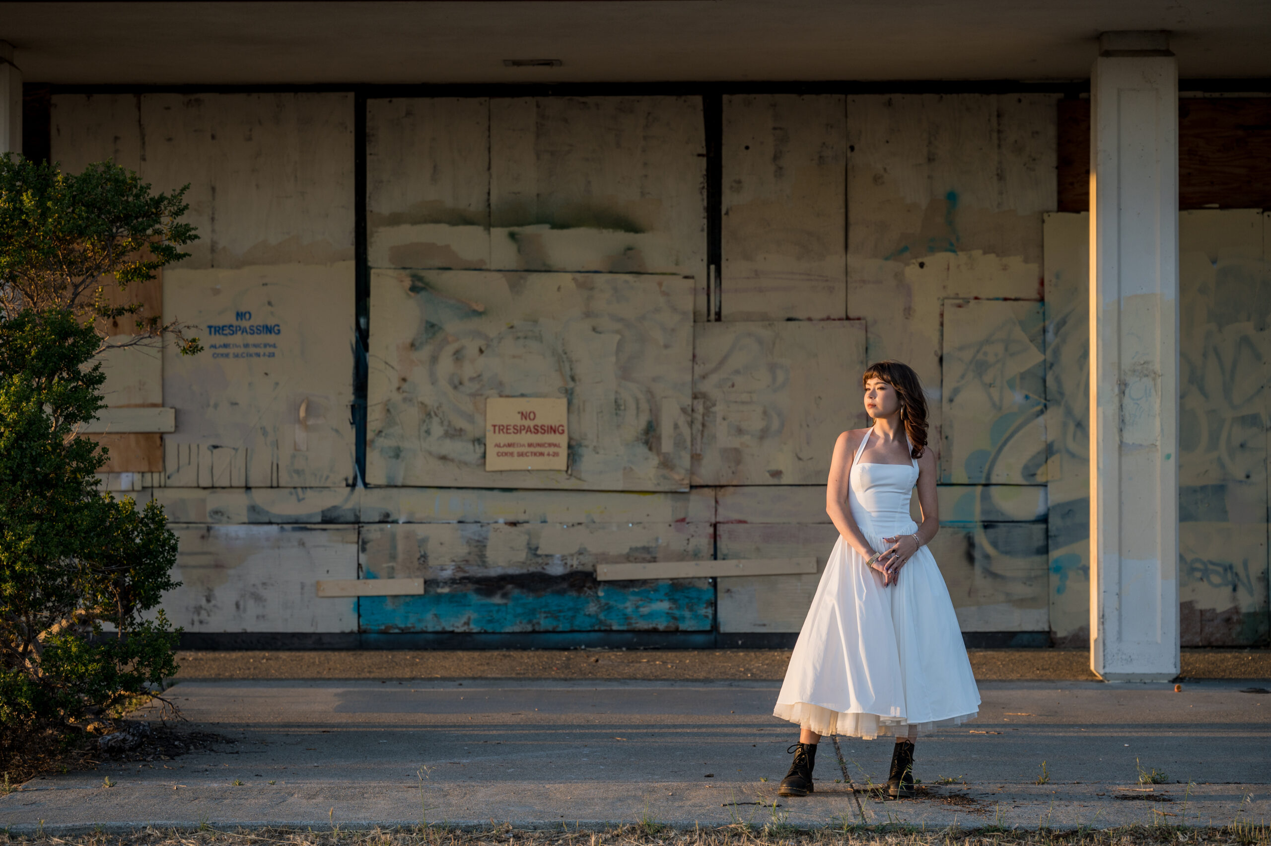 Golden hour senior portrait of a student in a white vintage-style dress and black boots standing in front of a boarded-up building with graffiti in Alameda.