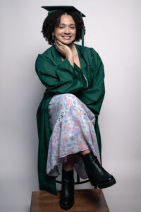 Studio senior portrait of a smiling young woman wearing a green graduation cap and gown, seated on a stool with a floral dress and black boots.