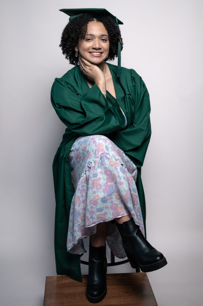 Studio senior portrait of a smiling young woman wearing a green graduation cap and gown, seated on a stool with a floral dress and black boots.