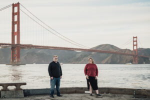 Outdoor senior photo with the Golden Gate Bridge in the background, featuring a student in a black sweater and jeans posing with a family member by the waterfront.