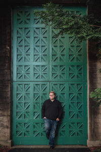 Bay Area senior portrait of a teen in casual jeans and a black sweater standing in front of a large green industrial door, framed by evergreen branches.