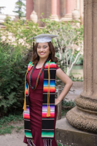 Bay Area senior photo of a graduate in a white cap and colorful stole, standing confidently in a burgundy dress outside the Palace of Fine Arts.