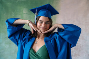 Close-up senior portrait of a graduate in a bright blue cap and gown, smiling and playfully framing their face with their hands in front of a soft green backdrop.