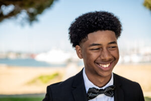 Candid senior photo of a young man in a black tuxedo and bowtie, smiling outdoors near the marina with soft-focus boats in the background.