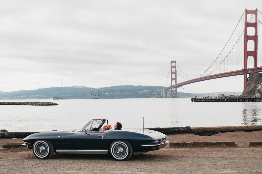 Woman wearing sunglasses and red scarf drives a vintage convertible past the Golden Gate Bridge in San Francisco on a cloudy day