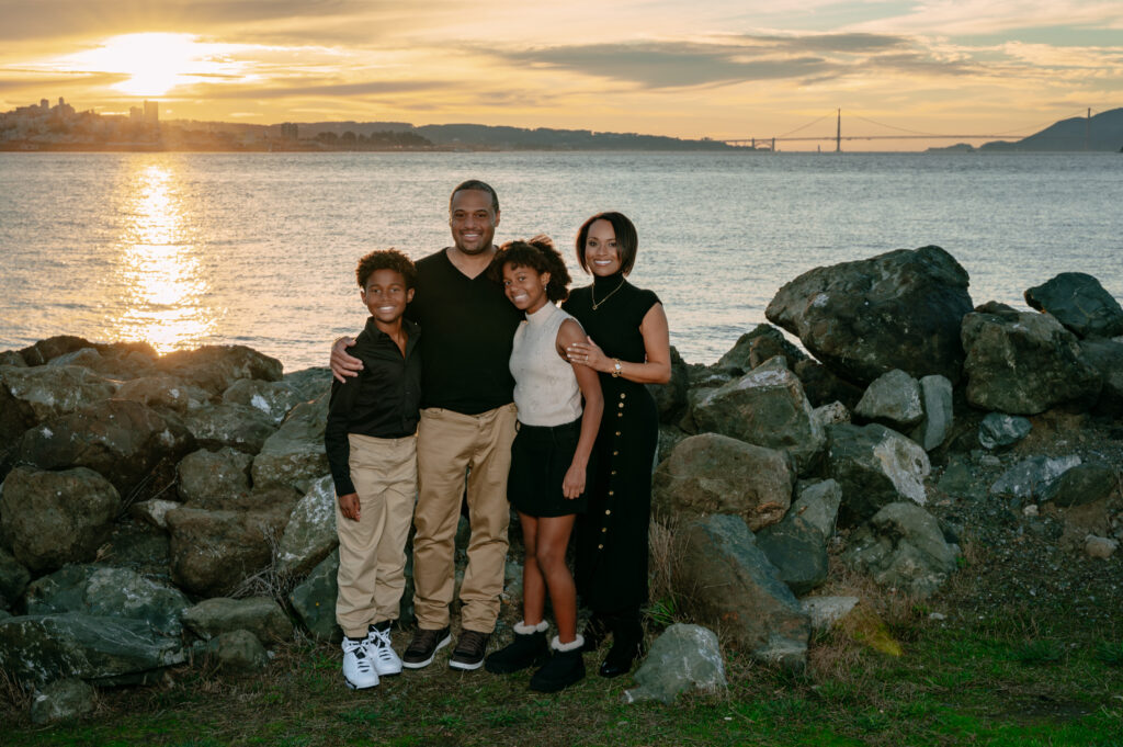 Family of five standing together near San Francisco Bay with Alcatraz and the Golden Gate Bridge in the distance, dressed in neutral tones